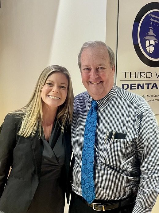 Smiling man in blue tie and woman with blonde hair standing in front of a dental office sign.