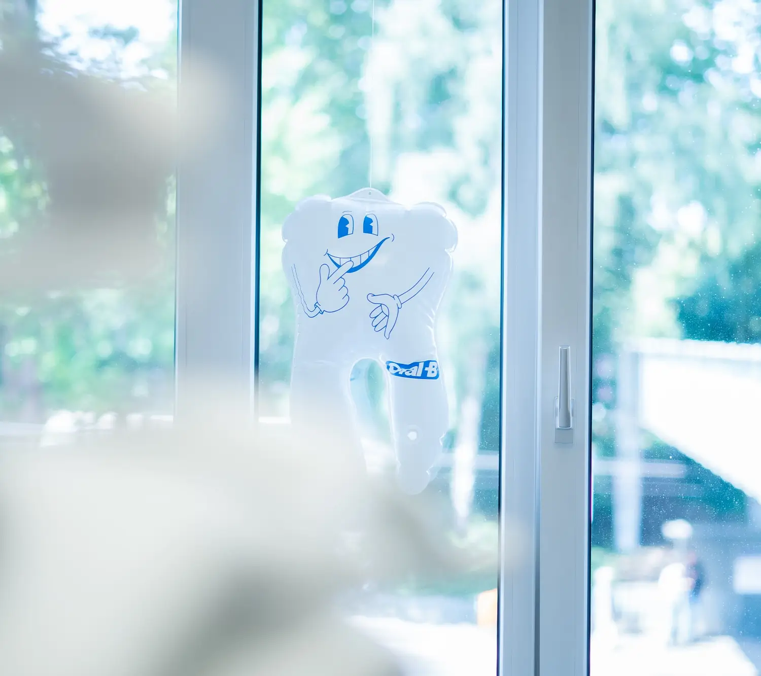 Weißer, zahnförmiger Ballon mit lächelndem Gesicht, der an einem Fenster hängt; unscharfer Vordergrund.