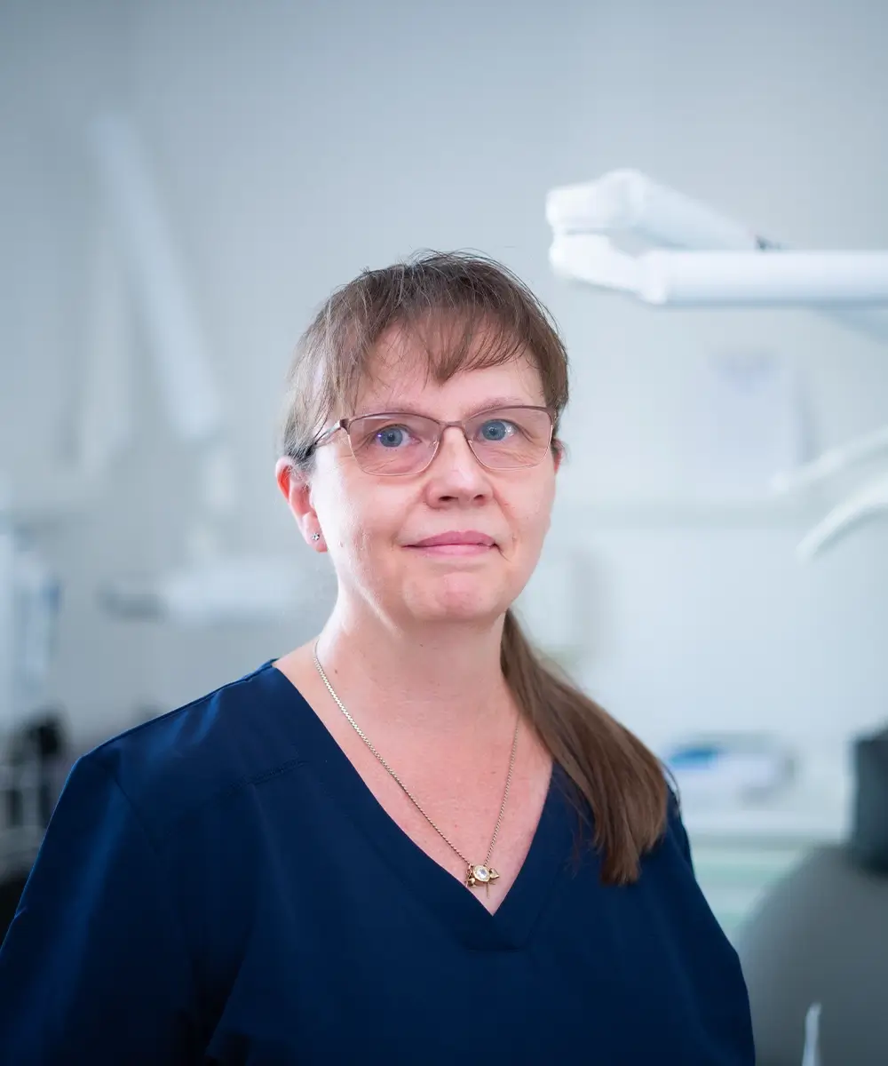 Eine Frau mit Brille in blauer Arbeitskleidung steht in einer Zahnarztpraxis; im Hintergrund sind unscharfe Geräte zu sehen.