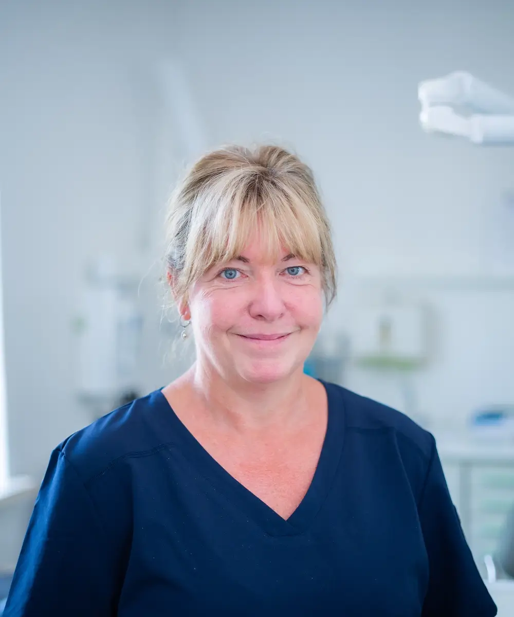 Lächelnde Frau mit blonden Haaren in einem dunkelblauen Kasack in einer klinischen Umgebung.