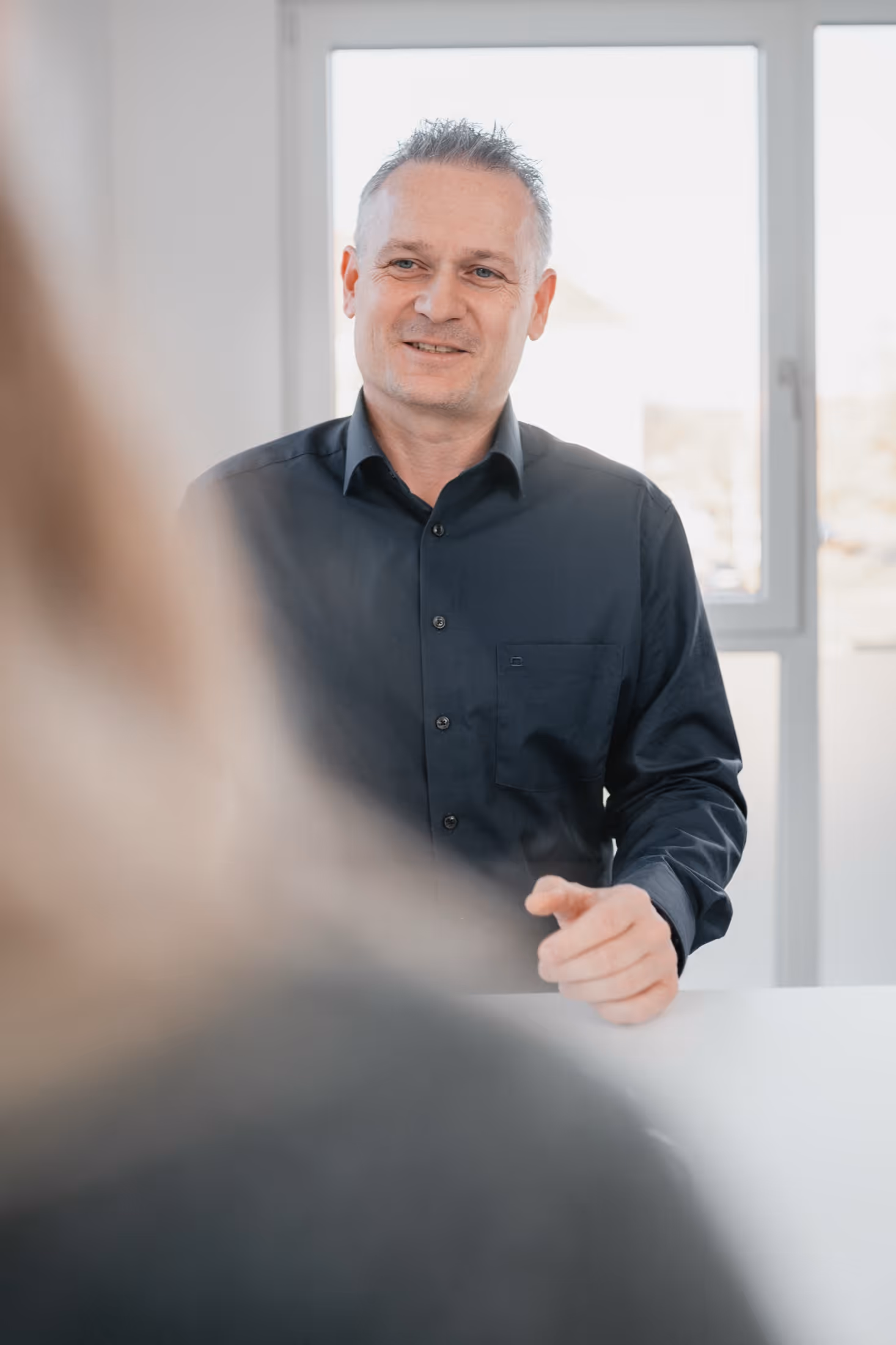 Mann im schwarzen Hemd spricht mit einer Person, die im Vordergrund unscharf ist.