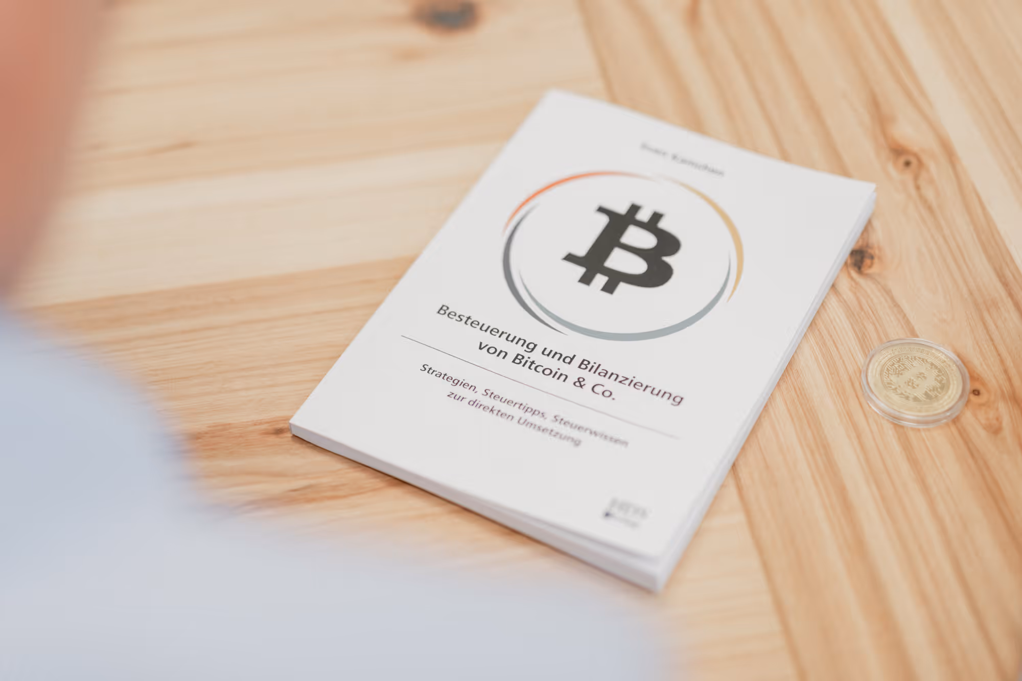 Buch über Besteuerung und Bilanzierung von Bitcoin liegt auf Holztisch neben Bitcoin-Münze.