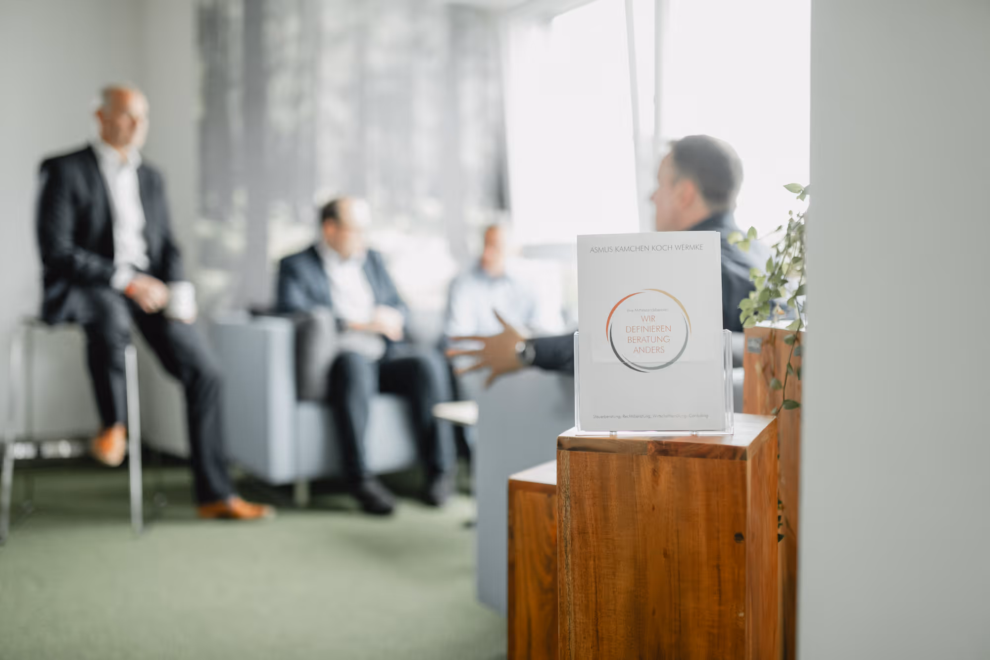 Vier Geschäftsmänner in Anzügen sprechen in einem modernen Büro mit grünem Teppich und Holzmöbeln.
