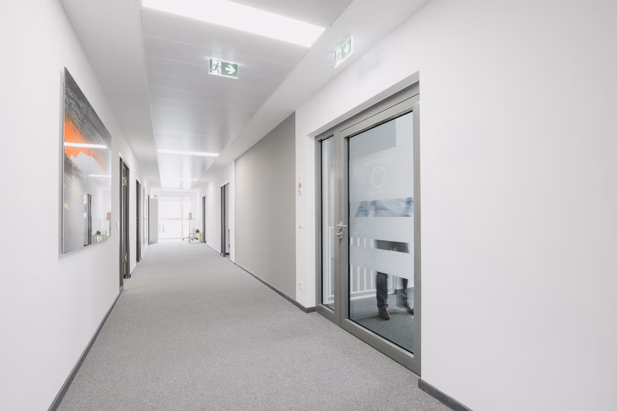 Heller Büroraumflur mit grauem Teppich, mehreren Türen und Glasschiebetür rechts.