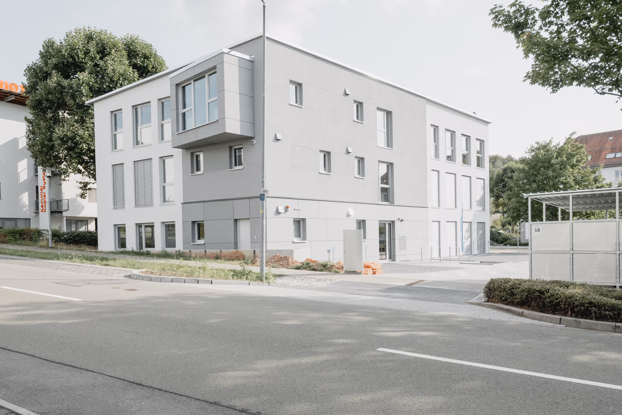 Modern, dreistöckiges graues Gebäude an einer Straße mit Bäumen und Parkplätzen.
