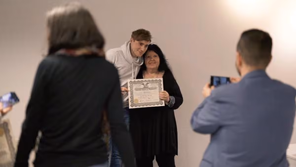 Junge und Frau posieren mit Diplom, zwei Personen fotografieren sie vor hellem Hintergrund.