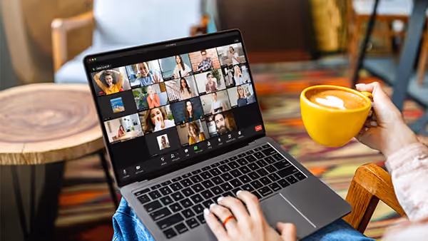 Person sitzt mit gelber Tasse in der Hand vor Laptop mit Videoanruf und mehreren Teilnehmern.