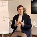 Junger Mann spricht gestikulierend vor Flipchart mit handgeschriebenen Notizen.