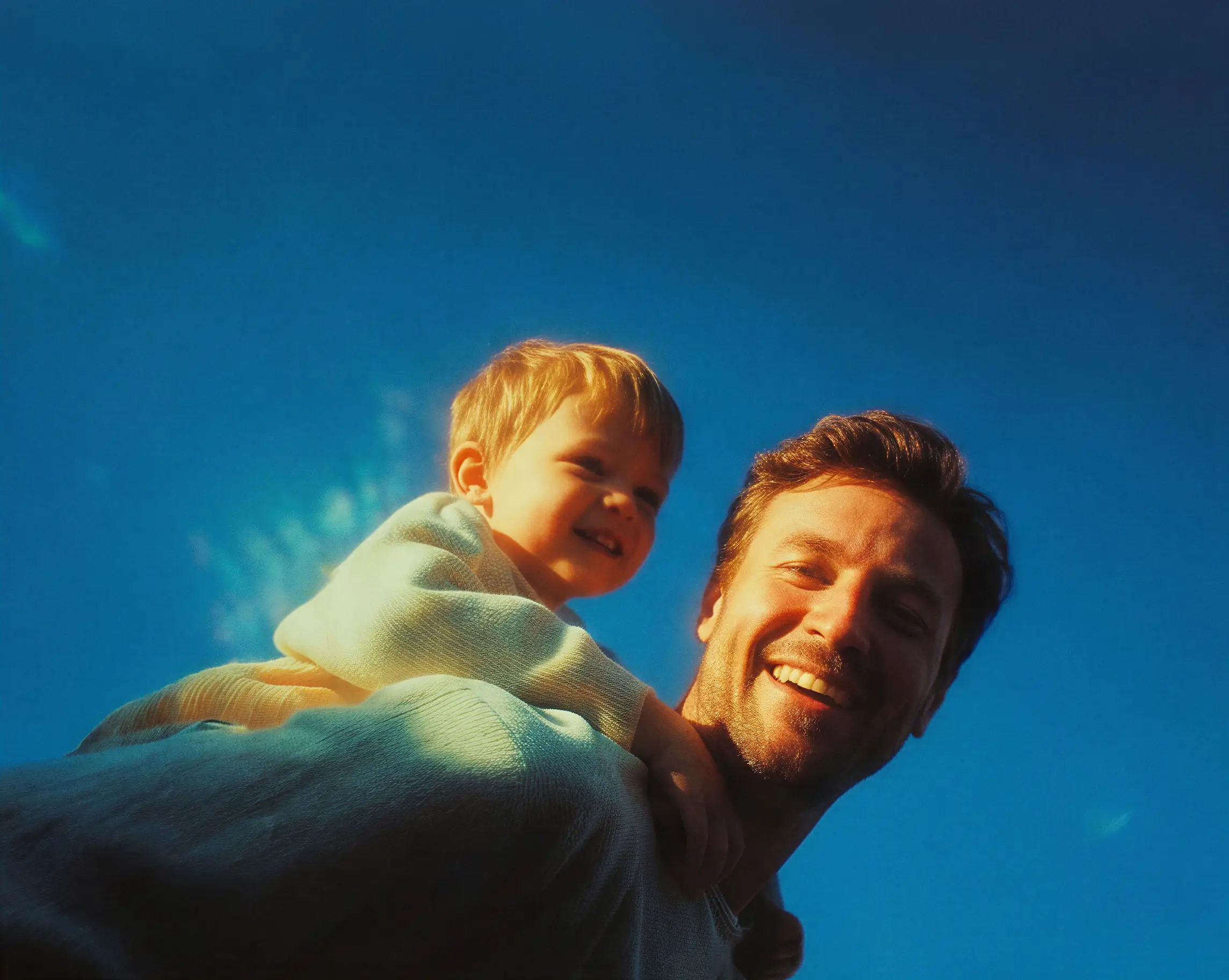 Smiling man giving a piggyback ride to a happy young child under clear blue sky.