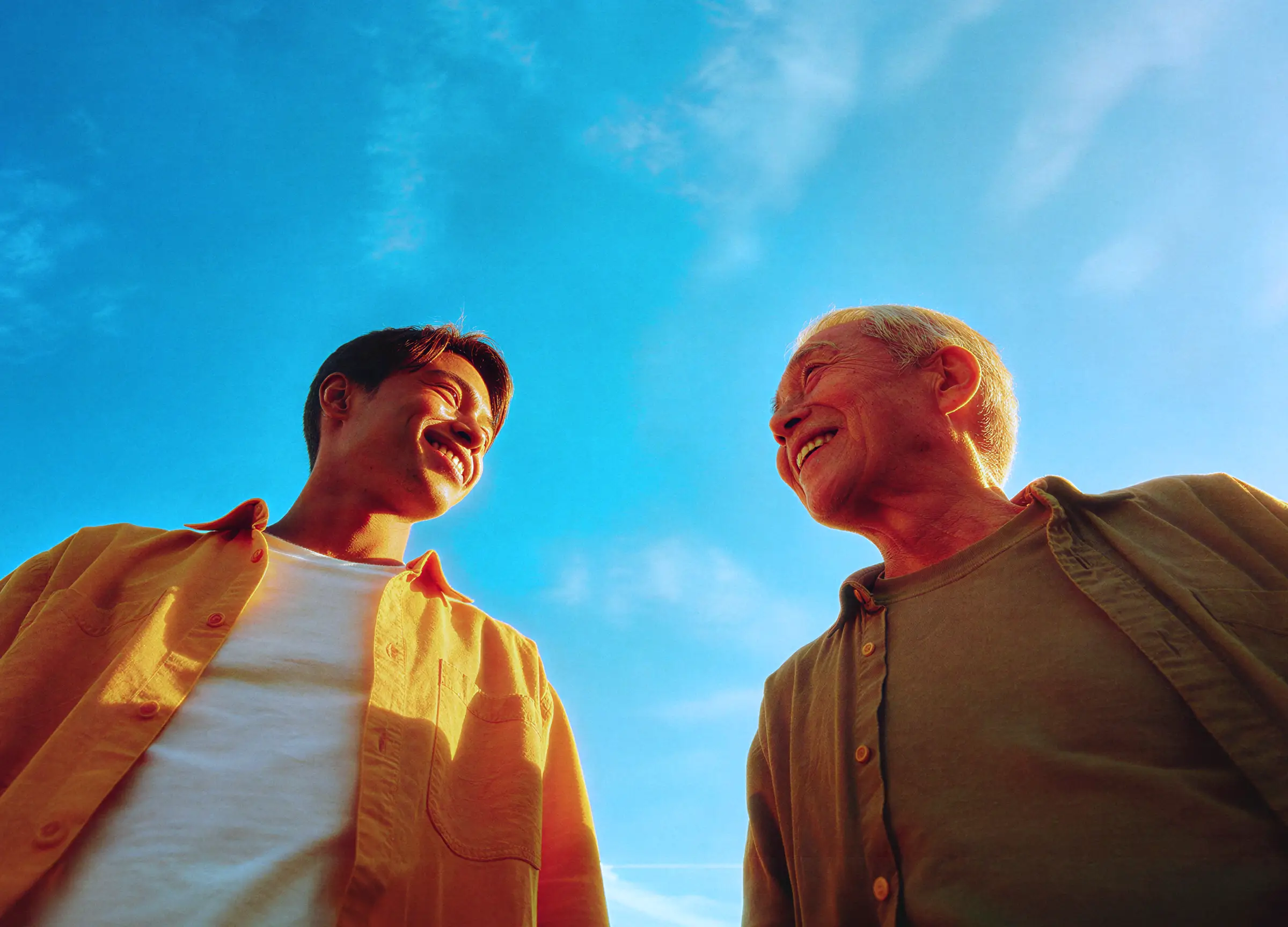 Young man and elderly man smiling at each other against a bright blue sky.