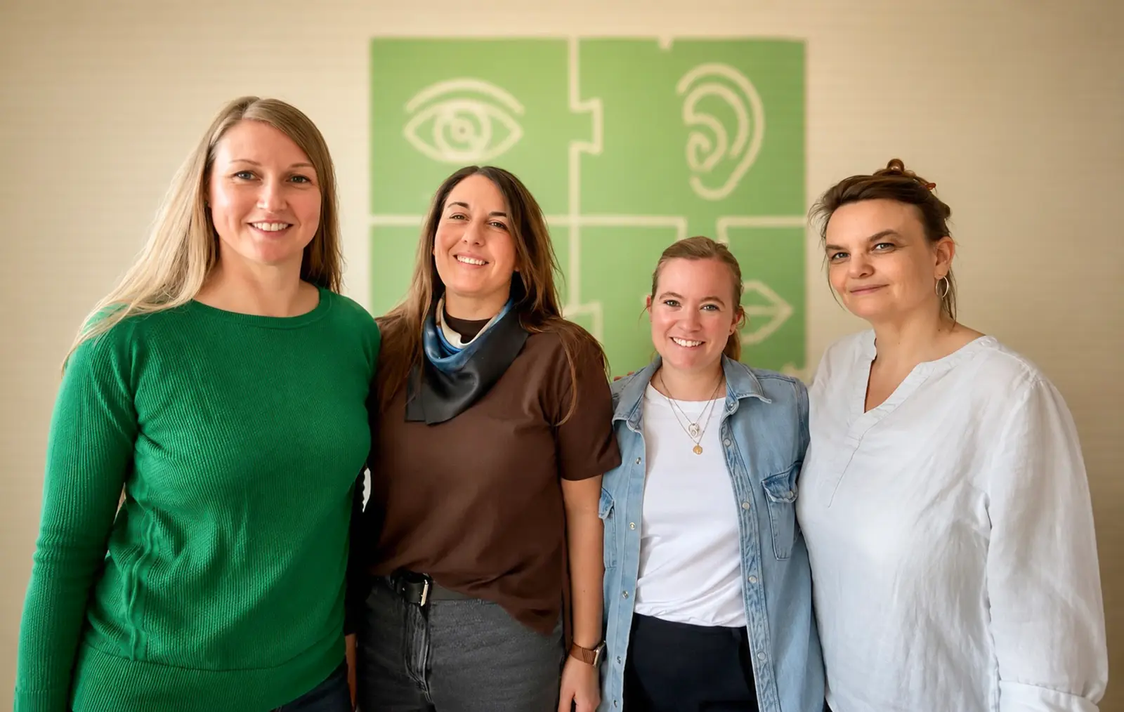 Elisabeth Warnke und ihr Team – vier lächelnde Frauen vor einer Wand mit grünen Puzzleteilen und Symbolen für Auge, Ohr und Mund.