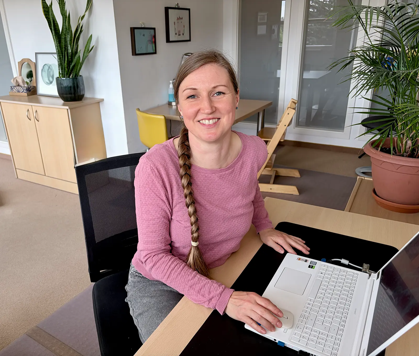 Elisabeth Warnke mit rosa Pullover und geflochtener Frisur sitzt lächelnd am Schreibtisch vor einem Laptop.
