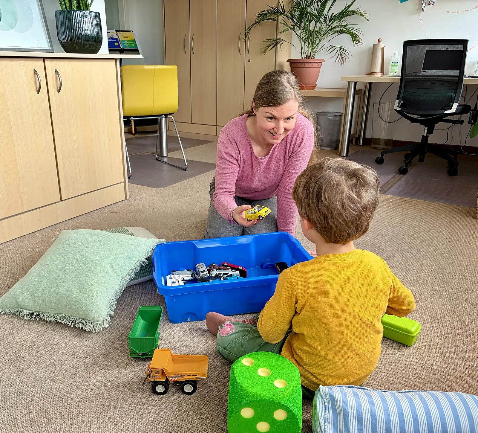 Frau spielt mit einem Jungen mit Spielzeugautos in einem Raum mit Teppich und Möbeln.