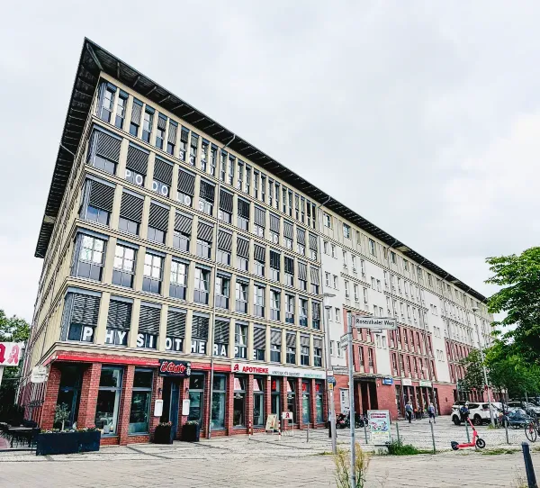 Mehrstöckiges Gebäude mit Ladenfronten und Gehweg unter bewölktem Himmel.
