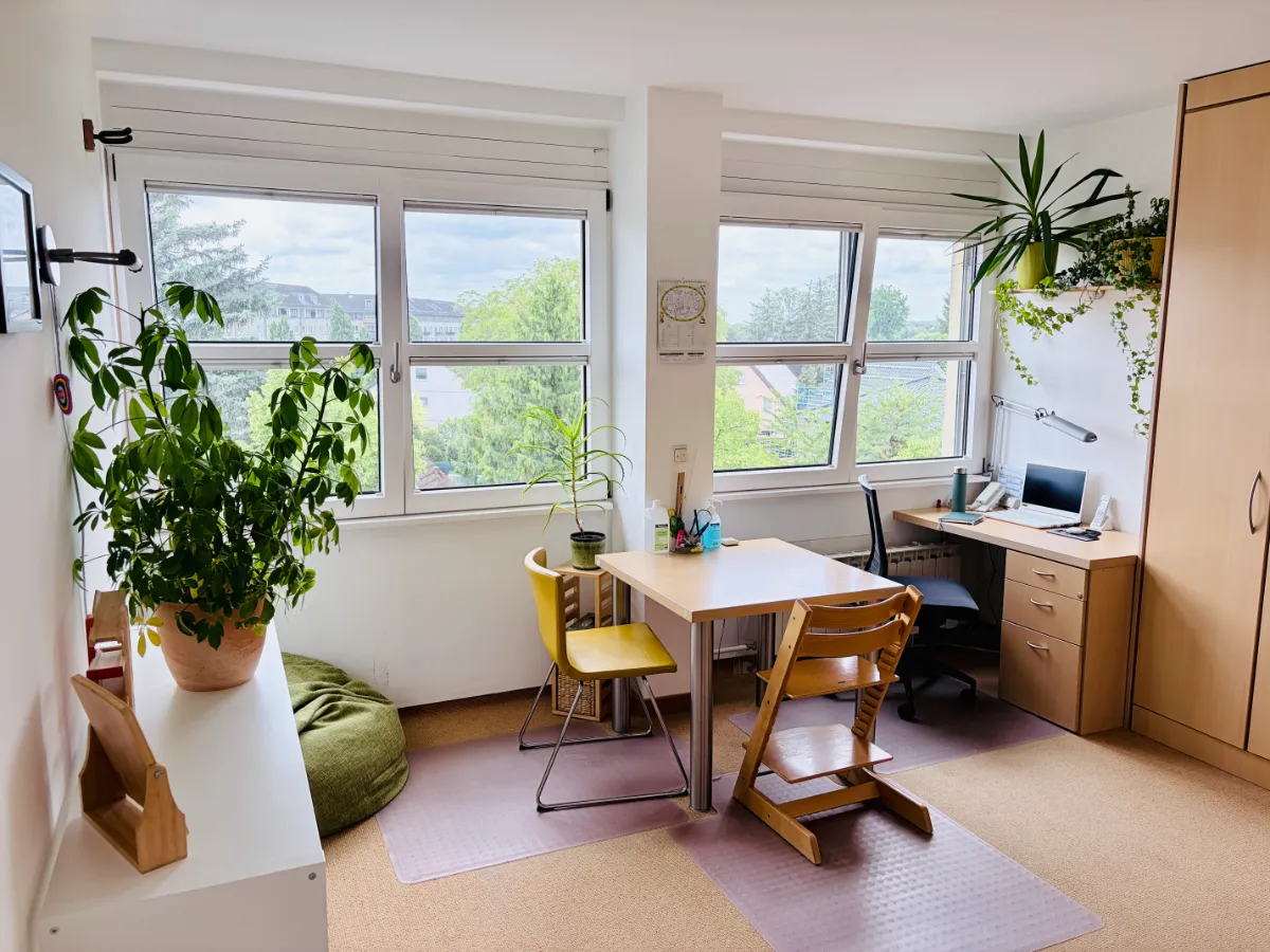 Helles Zimmer mit Tisch, verschiedenen Stühlen, Pflanzen, Schreibtisch und Fenster mit Blick ins Grüne.
