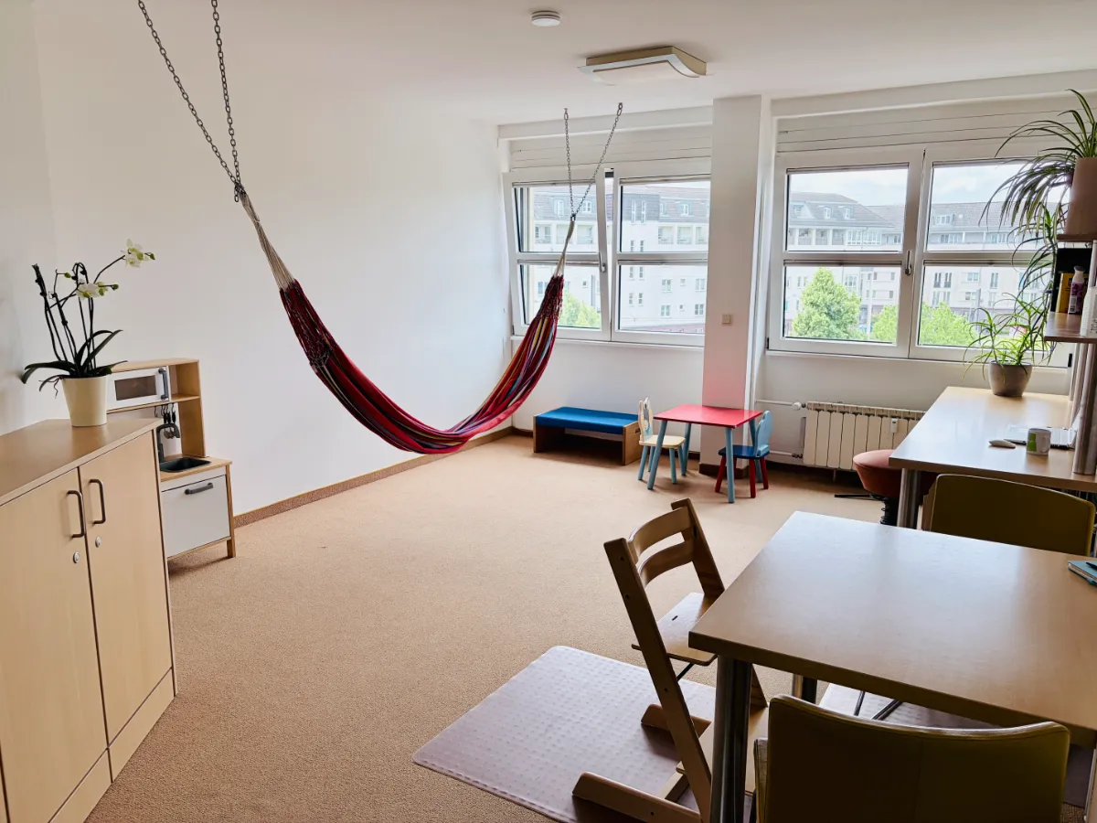 Heller Raum mit Hängematte, Kindertisch, Stühlen, Schränken und großen Fenstern.