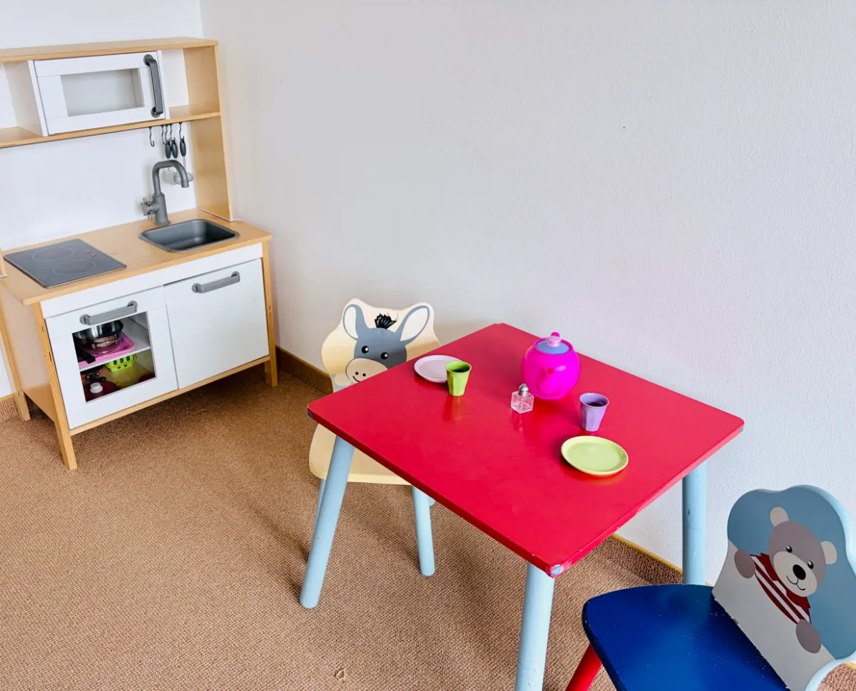 Kinderspielecke mit rotem Tisch, zwei bunten Stühlen und einer Spielküche an der Wand.