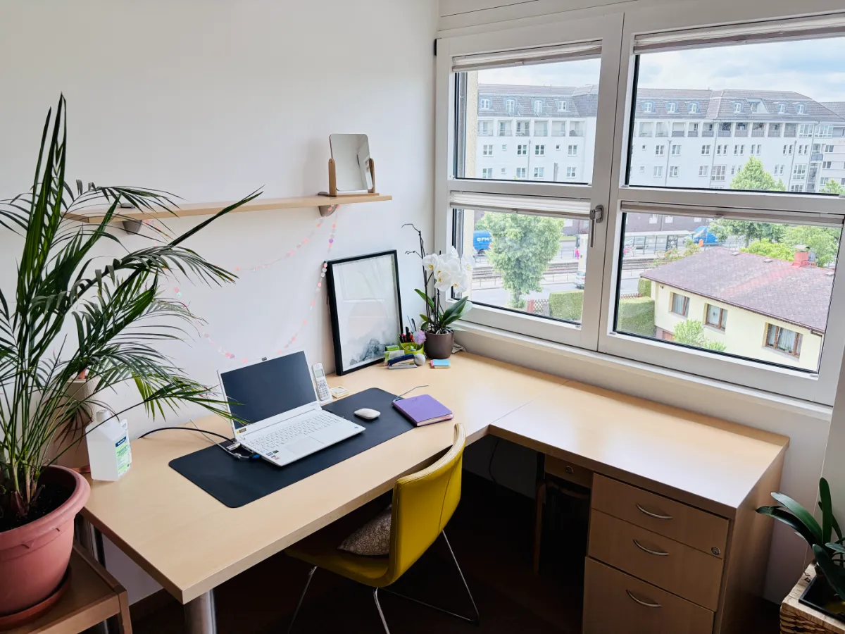 Helles Büro mit hellem Eckschreibtisch, Laptop, Pflanze und großem Fenster mit Blick auf Wohnhäuser.