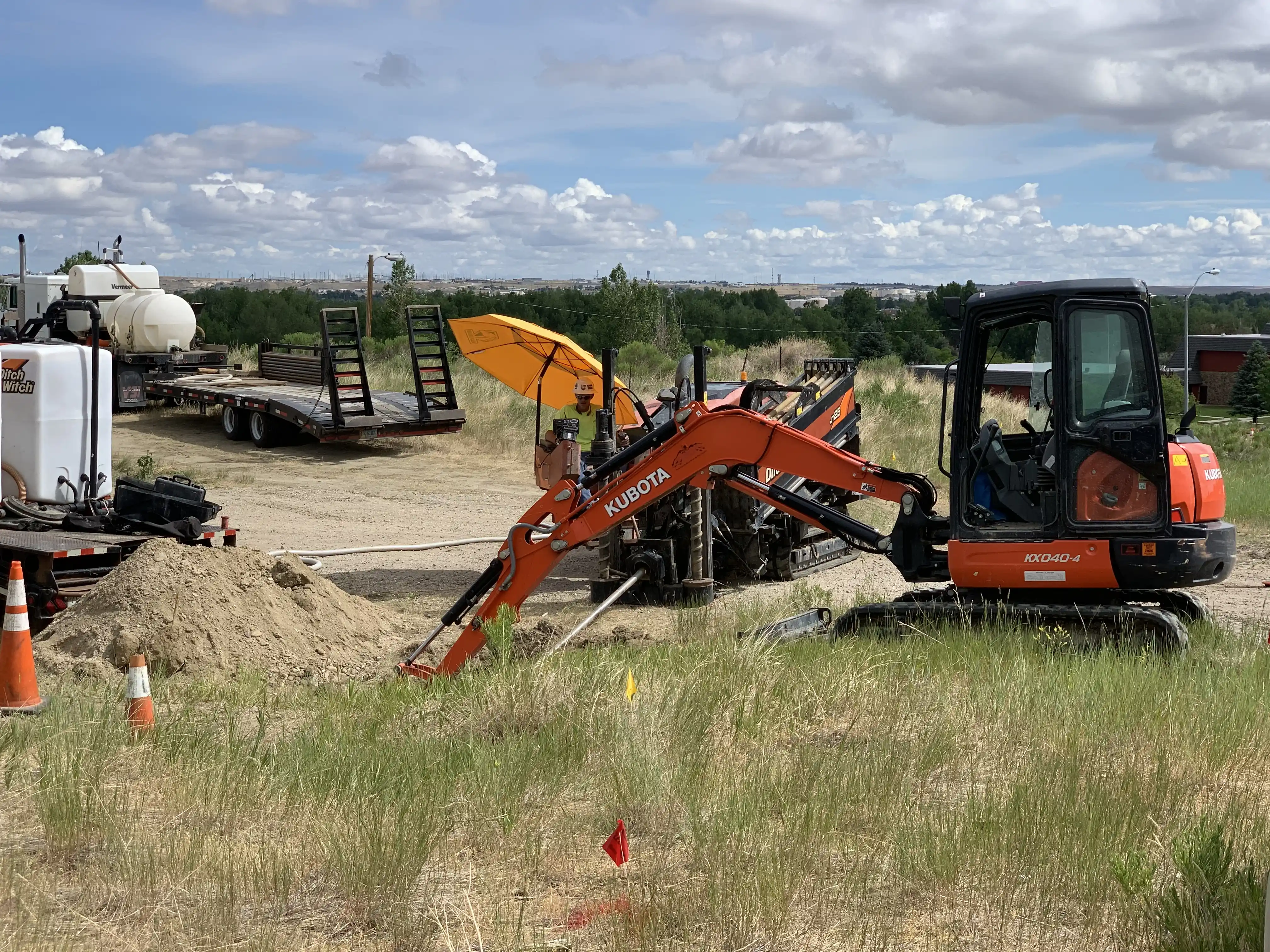 Orange Kubota excavator with extended arm at a construction site with dirt piles, traffic cones, and a man under a yellow umbrella.