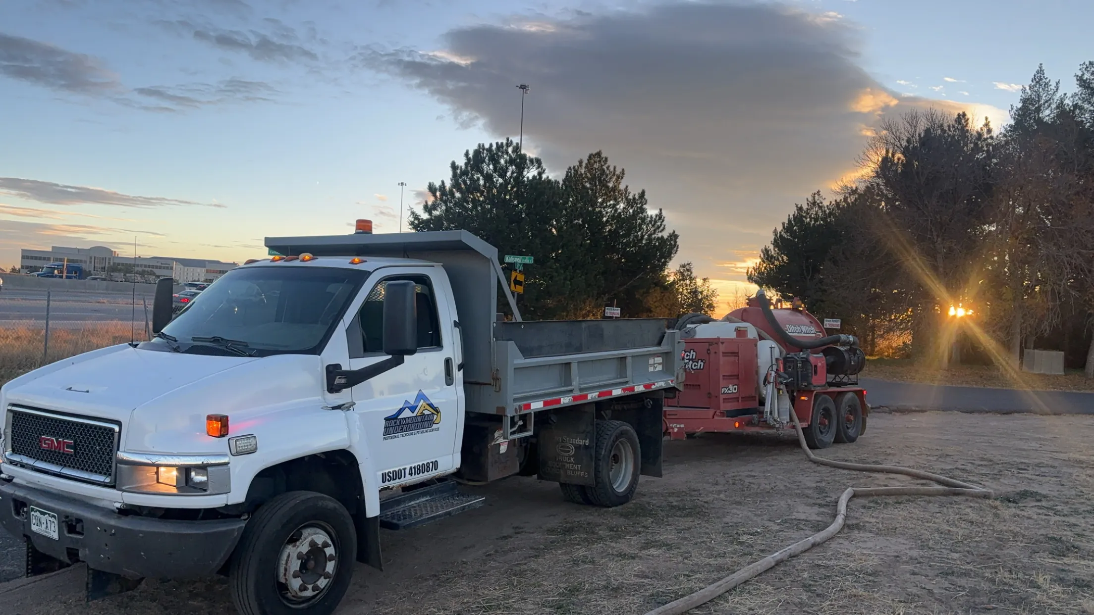 White GMC dump truck with Rocky Mountain Underground logo parked next to red Ditch Witch equipment at sunset.