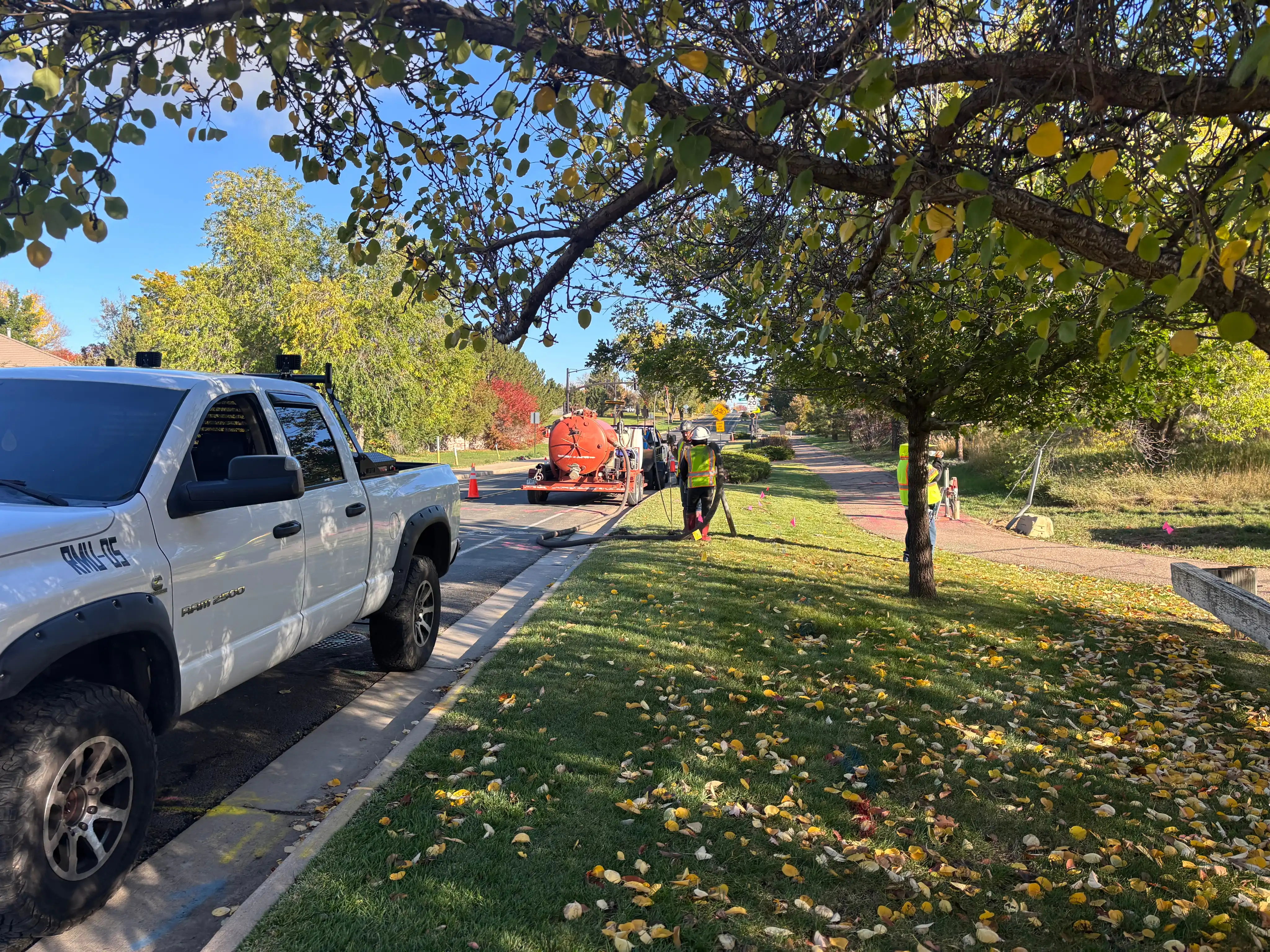 Workers in safety vests operating equipment near a white pickup truck on a suburban street lined with trees and fallen leaves.