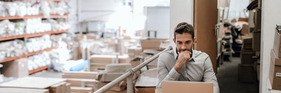 Warehouse manager reviewing cloud-based order management system on laptop amid inventory shelves and shipping boxes