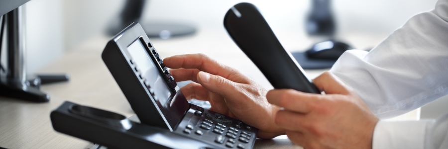 Business professional dialing a VoIP desk phone in an office, holding handset while pressing buttons on keypad