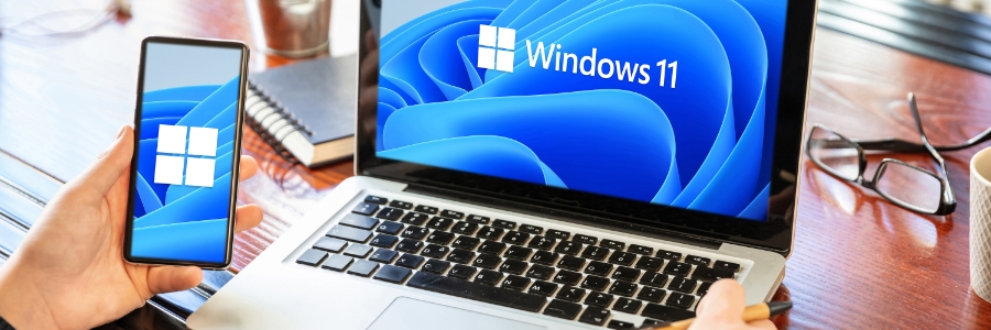Person holding smartphone displaying Windows 11 logo beside laptop with Windows 11 desktop on screen