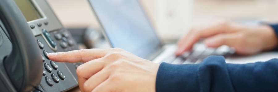 Person dialing a desk VoIP phone while typing on a laptop keyboard in an office environment