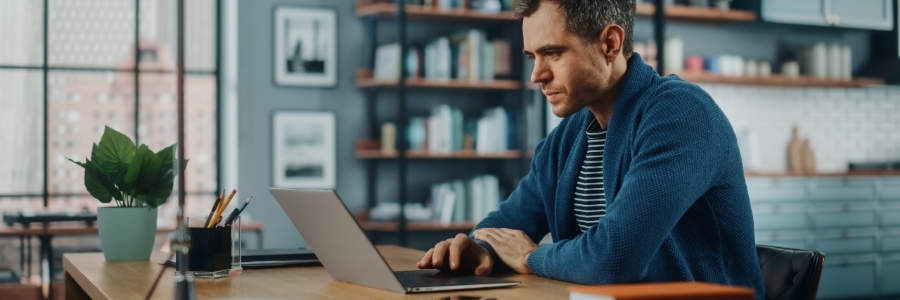 Man working remotely on laptop at home office desk with bookshelf background, using cloud-based productivity tools