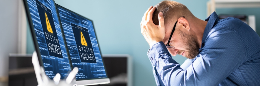 Stressed IT professional holding head in hands while dual monitors display system hacked cybersecurity warning alerts