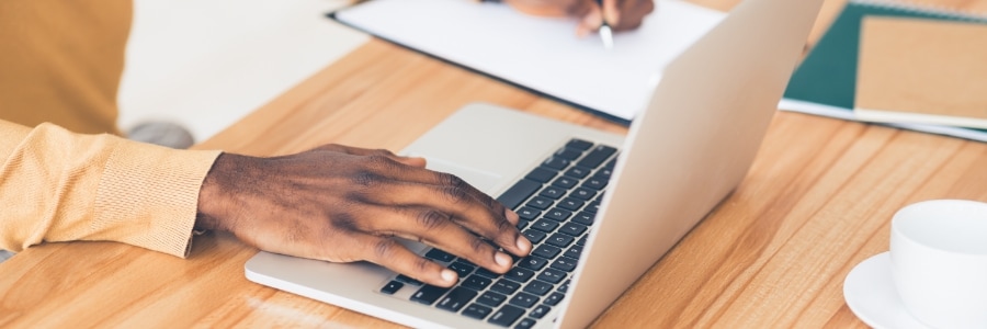 Person typing on laptop at wooden desk while colleague writes notes, representing IT support and computer maintenance