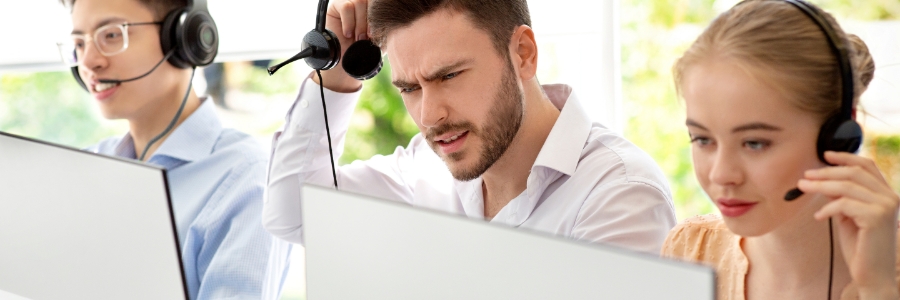 Three call center workers at computers with headsets, one visibly frustrated while troubleshooting a VoIP phone issue