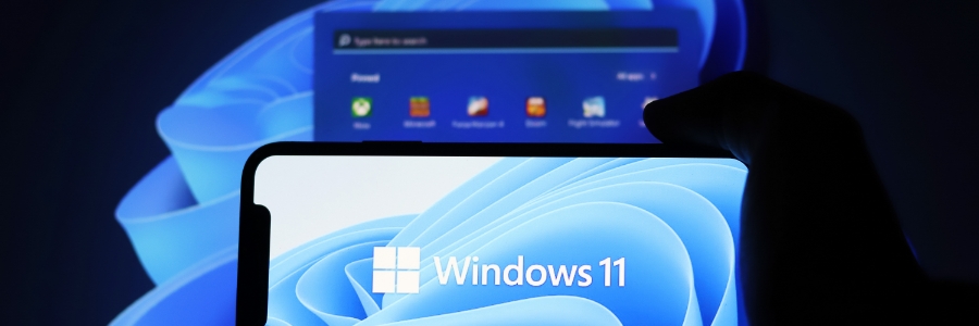 Hand holding smartphone displaying Windows 11 logo with desktop interface visible on monitor in background