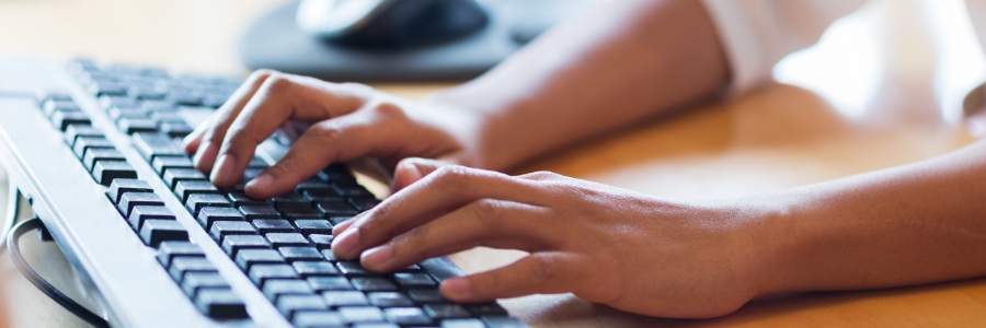 Hands typing on a black desktop computer keyboard with a mouse in the background on a wooden desk