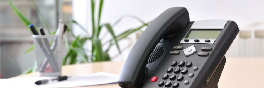 Black VoIP desk phone on office desk with pen holder and green plant in background