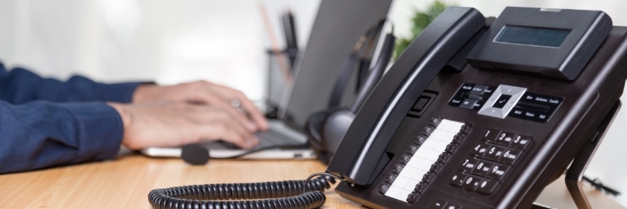 Business desk with VoIP office phone in foreground and person typing on laptop in background