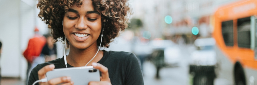 Woman smiling while using smartphone with earbuds on a city street, exploring business communication solutions