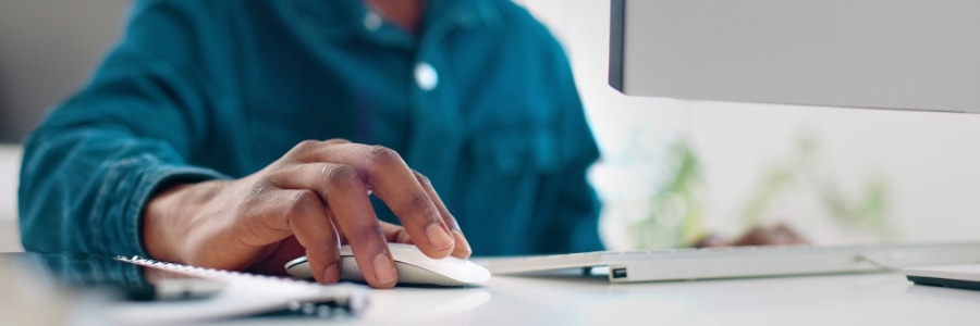 Person using a white computer mouse at a desk with a keyboard and monitor for office IT work
