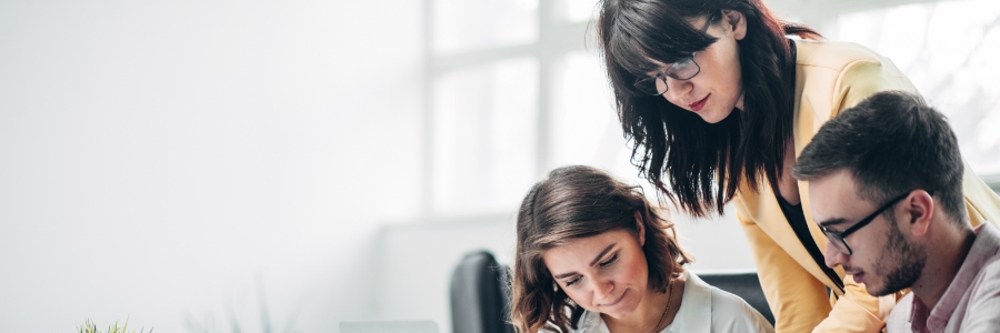 IT professional standing over two colleagues reviewing work on a laptop in a modern office