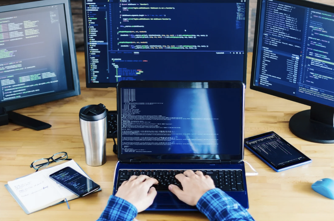 IT professional typing on laptop surrounded by multiple monitors displaying code and terminal output