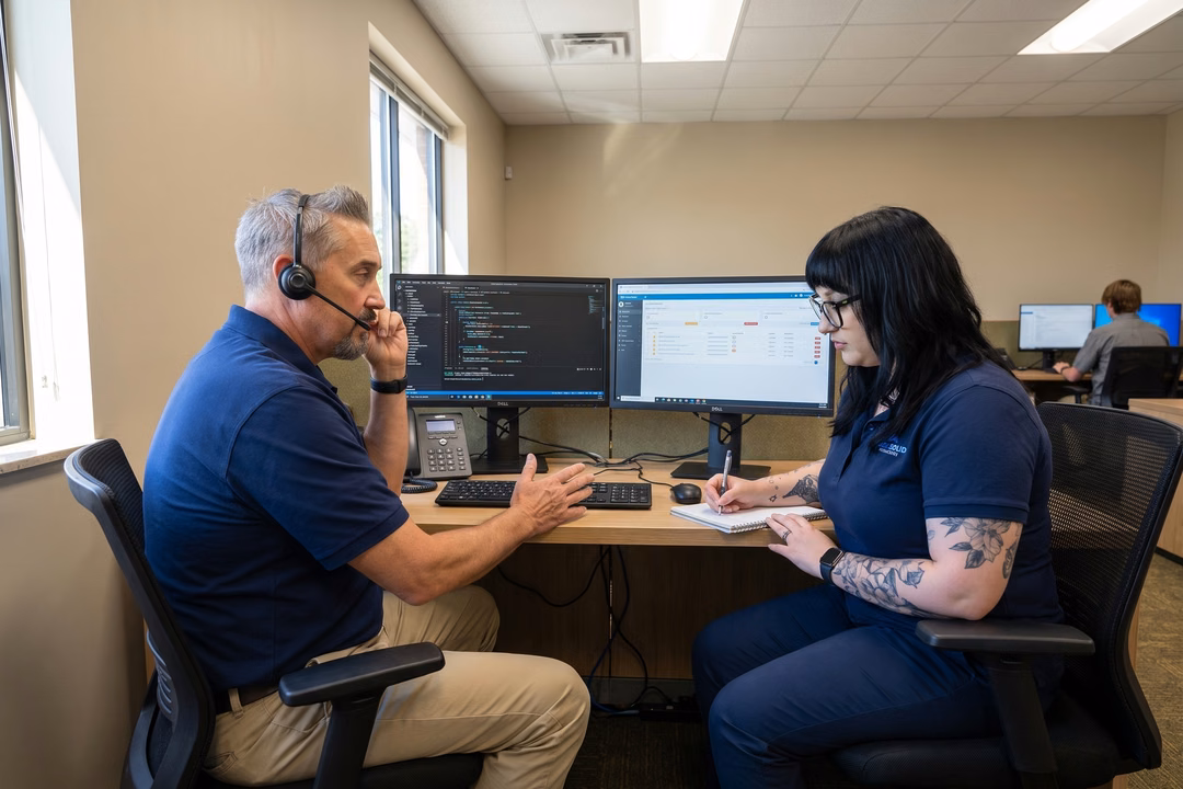 IT support technician with headset consulting with colleague taking notes at dual-monitor workstation in Thousand Oaks office