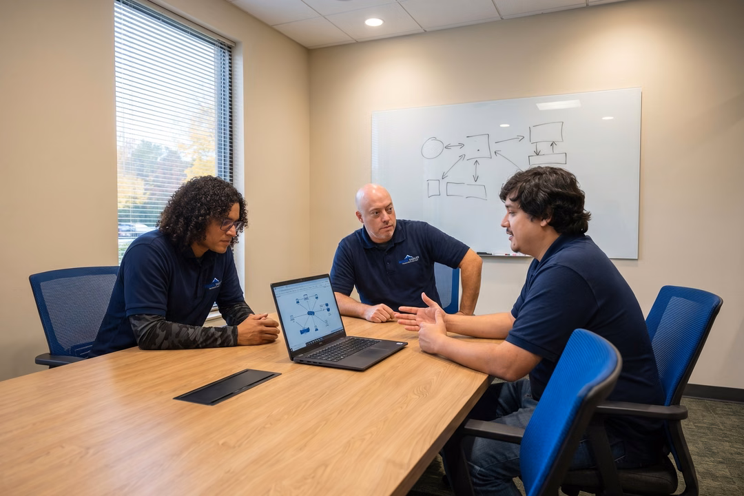 Three IT professionals reviewing cloud infrastructure diagrams on a laptop in a conference room with a whiteboard