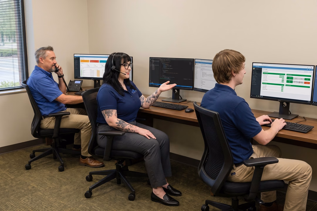 IT support team working at computers, one speaking on phone, one wearing headset, one typing, in a modern office