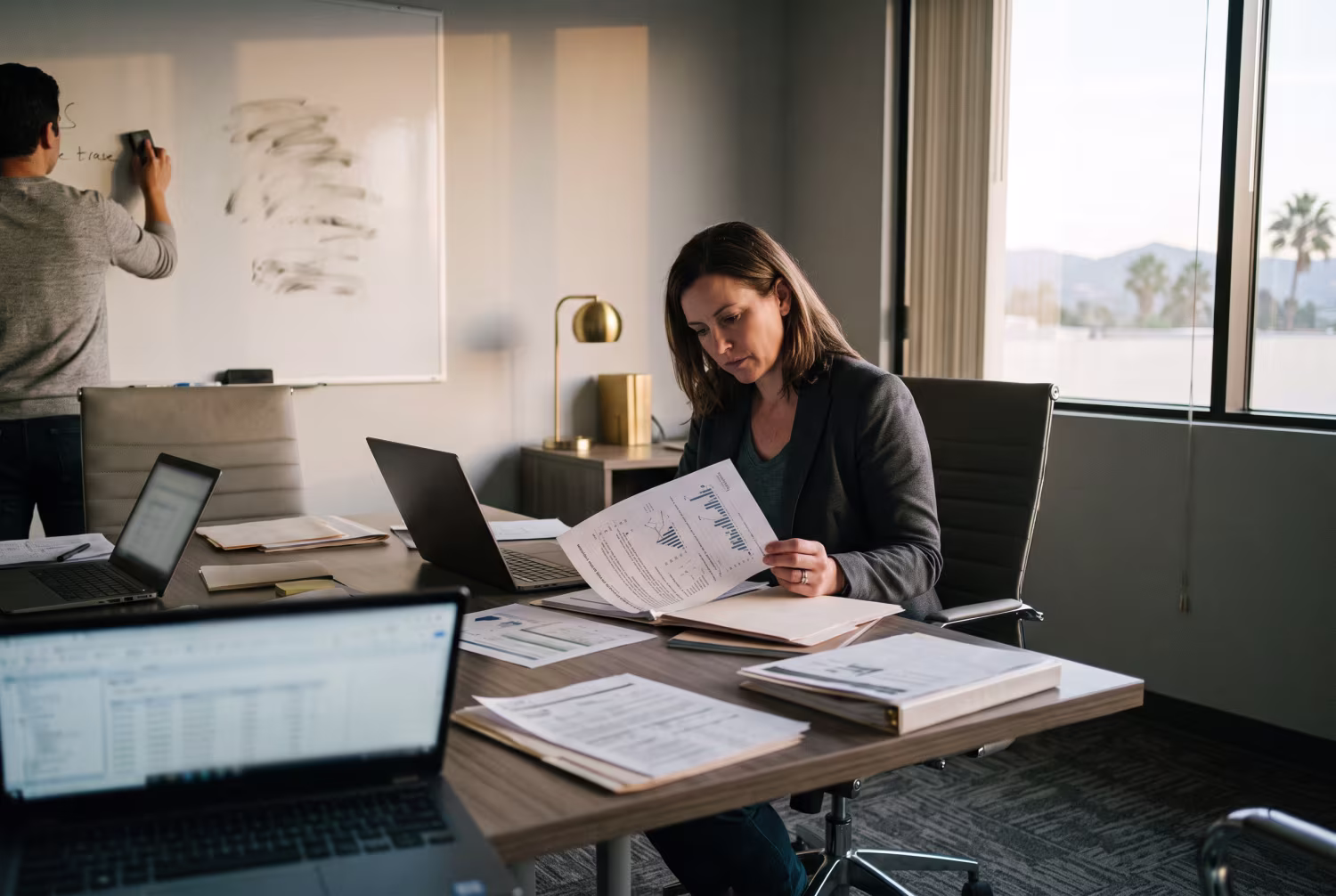 IT manager studying financial report at table, colleague at whiteboard, California office scene