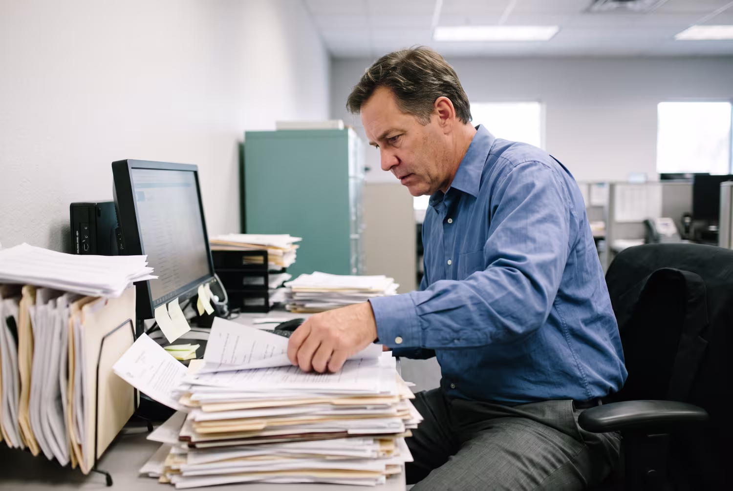 Mid-aged man sorting paper documents, idle computer implying SharePoint underutilization