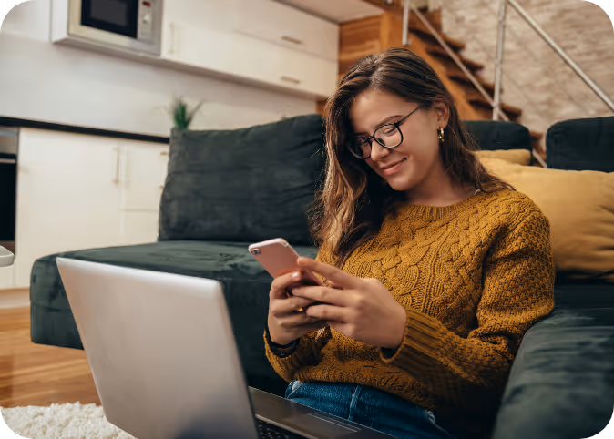 Smiling woman wearing glasses and a mustard sweater using a smartphone with a laptop on her lap in a modern living room.