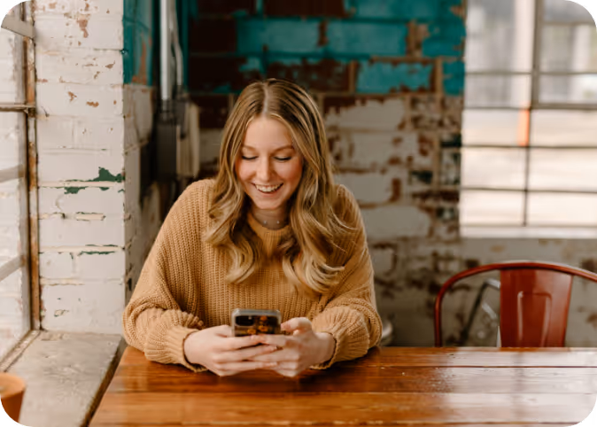 Smiling young woman with long wavy hair in a tan sweater looking at her phone at a wooden table.