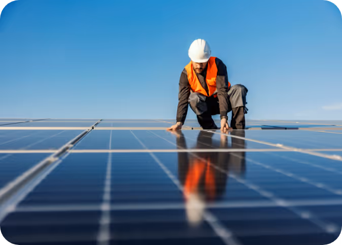 Worker wearing a white helmet and orange vest inspecting solar panels under a clear blue sky.