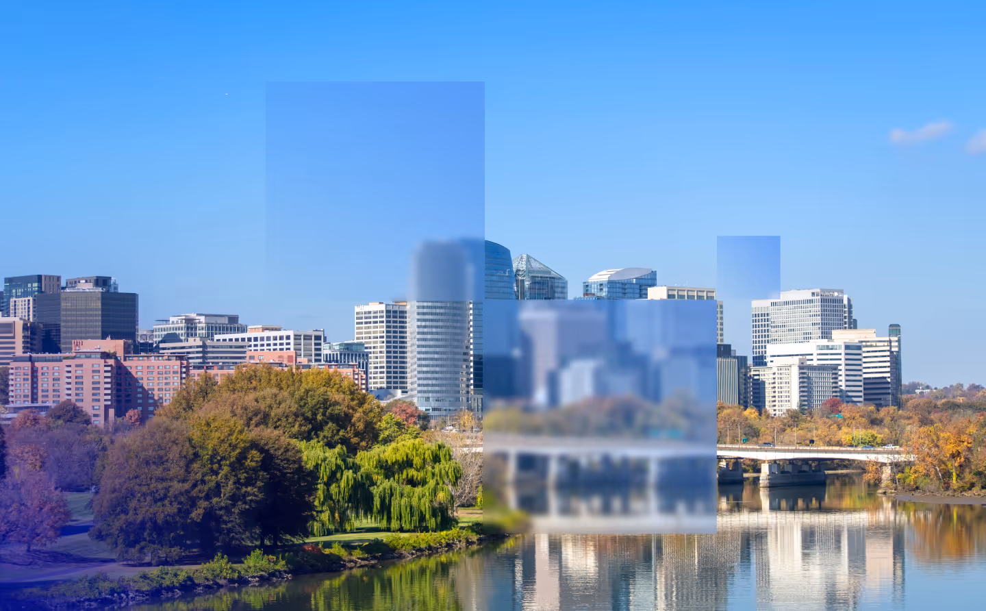 City skyline with modern buildings, trees, and a river reflecting structures on a clear sunny day.